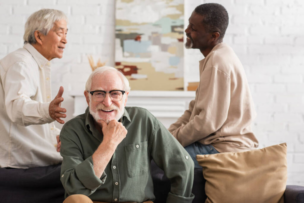Positive elderly man looking at camera near interracial friends at home  - Foto, imagen