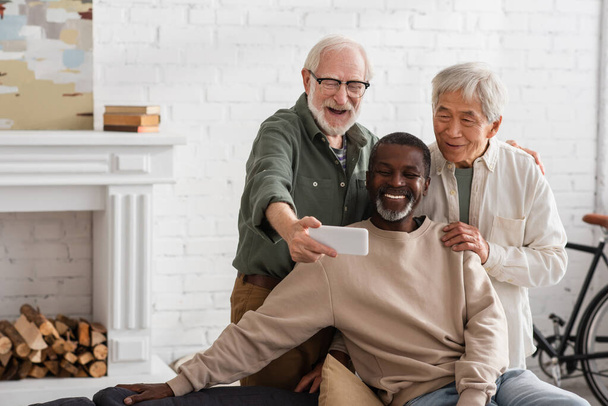 Elderly man taking selfie on smartphone near multiethnic friends at home  - Photo, Image