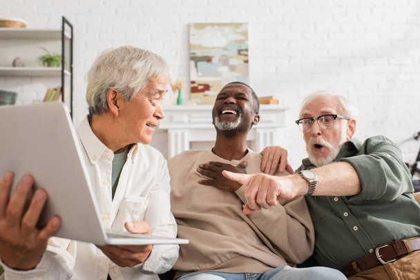Positive asian man holding laptop near interracial friends at home  - Photo, Image