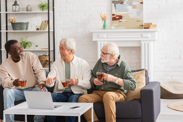 Smiling asian man with tea showing like near multiethnic friends and devices at home  - Photo, Image