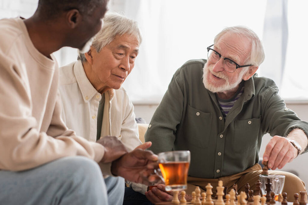 Smiling man playing chess with interracial friends near tea at home  - Photo, image