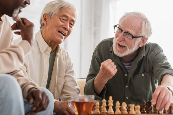 Excited senior man playing chess with multiethnic friends at home  - Foto, Imagen