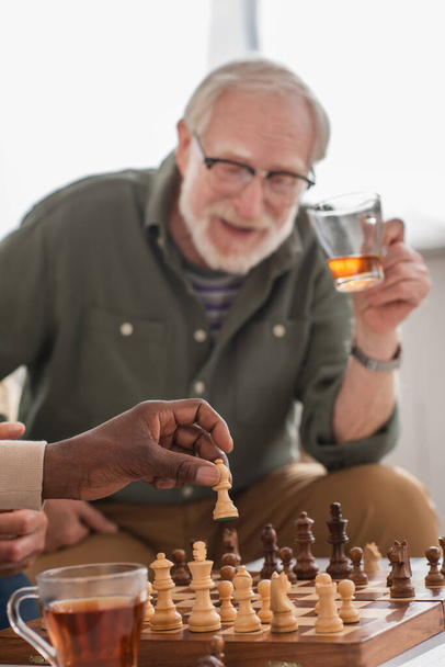 African american man holding chess figure near tea and blurred friends at home  - Valokuva, kuva