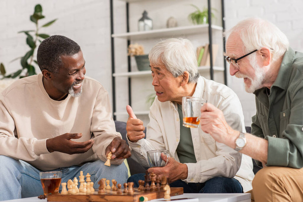 Asian man holding tea and showing like sign near multiethnic friends and chess at home  - Photo, image