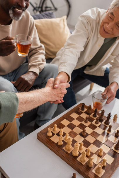 Vista de ángulo alto de amigos interracial estrechando las manos cerca del hombre afroamericano con té y tablero de ajedrez  - Foto, Imagen