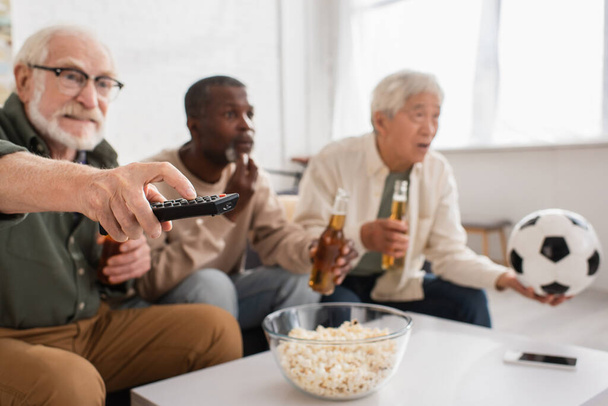 Hombre mayor sosteniendo mando a distancia cerca borrosa interracial amigos con cerveza en casa  - Foto, Imagen