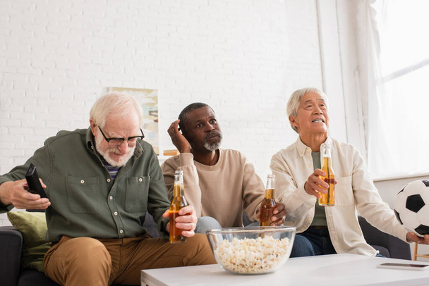 Stressed interracial pensioners holding beer and football near popcorn in living room  - Foto, imagen