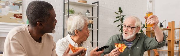 Старший мужчина держит пиццу и пиво возле мультикультурных друзей с пультом дистанционного управления на дому, баннер  - Фото, изображение