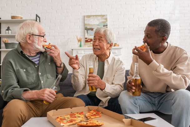 Starszy azjata mężczyzna trzyma piwo i wskazuje palcem blisko międzyrasowych przyjaciół jedzących pizzę w domu  - Zdjęcie, obraz