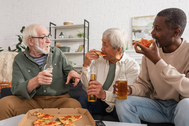 Elderly man holding beer bottle and talking to interracial friends eating pizza in living room  - Valokuva, kuva