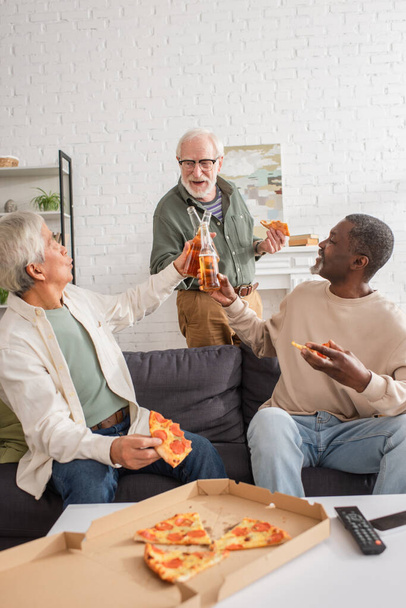Amigos mayores multiculturales que tintinean cerveza cerca de sabrosa pizza y controlador remoto en casa  - Foto, Imagen