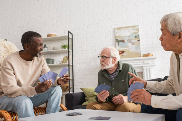 Sonriente hombre afroamericano sosteniendo cartas cerca de amigos multiétnicos en la sala de estar  - Foto, imagen