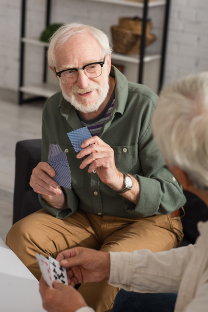 Homme âgé souriant tenant des cartes à jouer près d'un ami flou à la maison  - Photo, image
