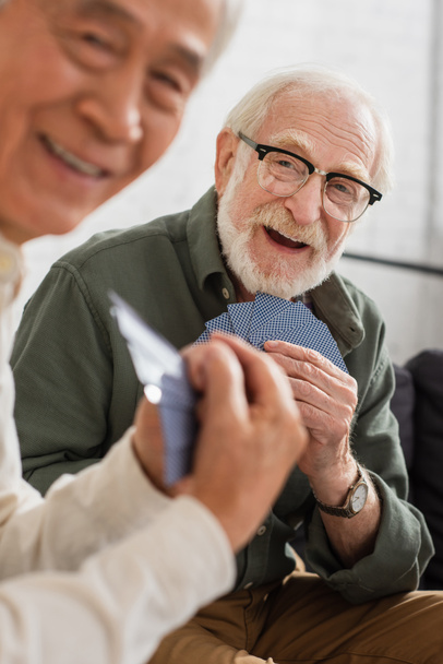 Smiling grey haired man holding playing cards near blurred asian friend at home  - Photo, Image