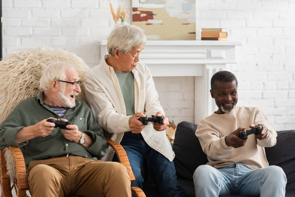 KYIV, UKRAINE - OCTOBER 12, 2021: Smiling african american man playing video game near multiethnic friends at home  - Fotografie, Obrázek