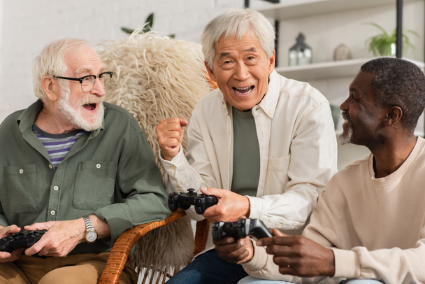 KYIV, UKRAINE - OCTOBER 12, 2021: Excited asian man showing yes gesture while playing video game with multiethnic friends at home  - Fotografie, Obrázek