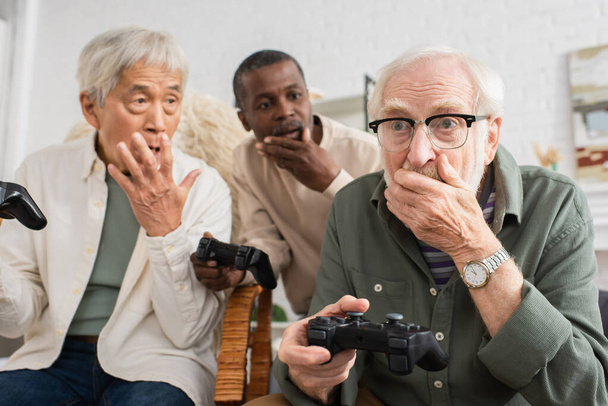 KYIV, UKRAINE - OCTOBER 12, 2021: Shocked senior man playing video game with blurred multicultural friends at home  - Foto, Imagem