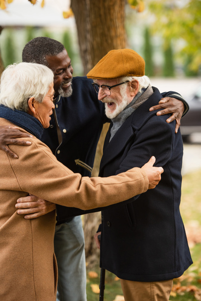 Cheerful multiethnic senior men hugging in park in autumn  - Photo, image