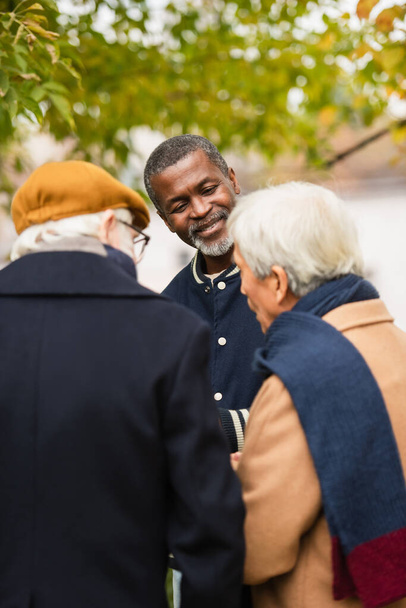 Sonriente hombre afroamericano mirando borrosa amigos multiétnicos en el parque  - Foto, imagen