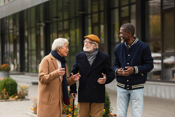 Positive senior multiethnic men talking while walking on urban street  - Foto, imagen