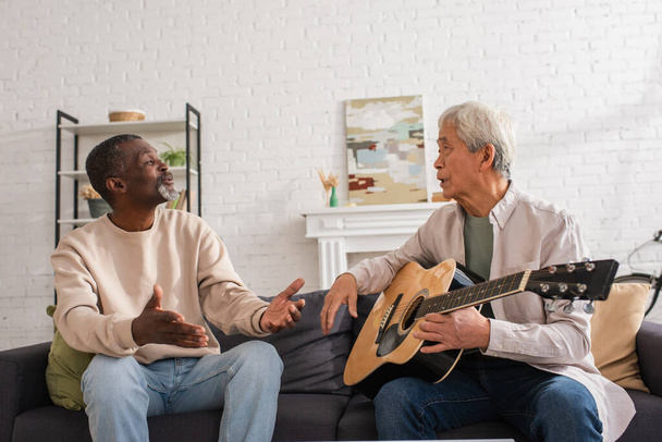 Gris peludo asiático hombre celebración acústica guitarra cerca africano americano amigo en casa  - Foto, imagen