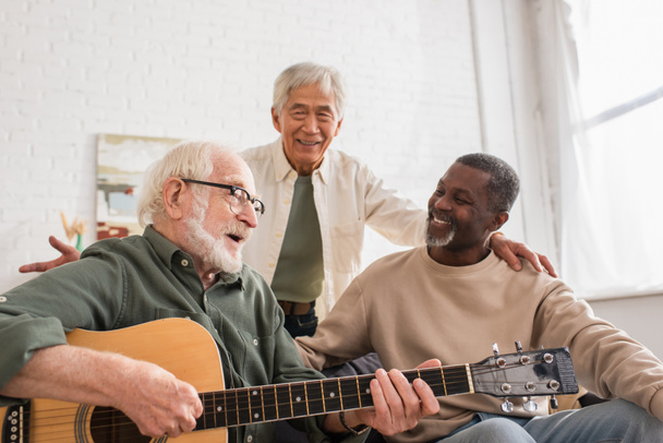Grey haired man playing acoustic guitar near interracial friends at home  - Foto, Imagem