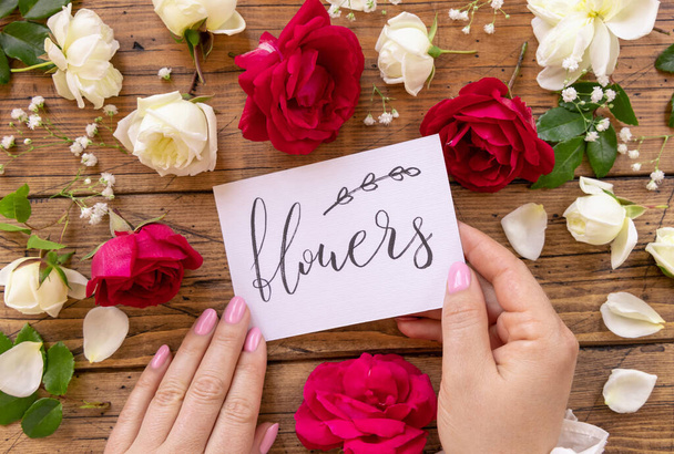 Hands with handwritten card FLOWERS surrounded by red and cream roses close up on a wooden table. Femminine romantic declaration of love near flowers, Valentines day concept - Photo, Image