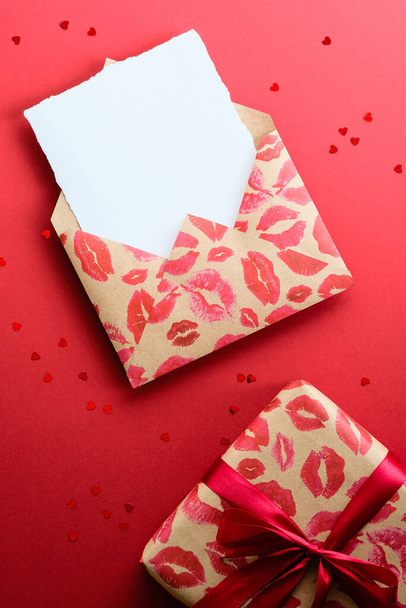 Craft paper envelope with lipsticks marks and gift box on red background with heart shaped confetti. Happy Valentine's Day concept. Flat lay, top view. - Φωτογραφία, εικόνα