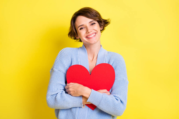 Photo of young cheerful girl happy positive smile hug big red heart feeling romantic isolated over yellow color background - Фото, изображение