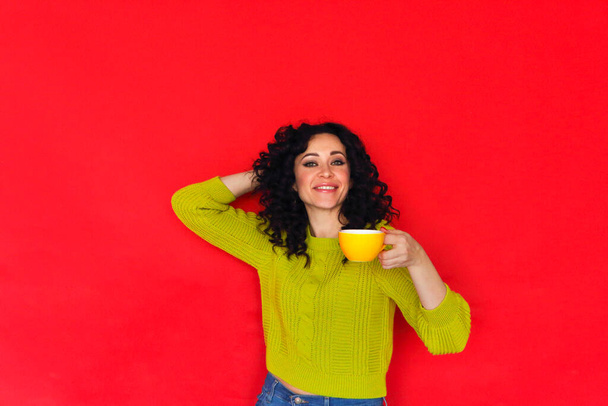 Joven mujer rizada feliz sosteniendo la taza de té, posando aislado en la pared roja. Sonriendo hembra satisfecha disfrutando de la bebida caliente, bebiendo café de la taza mientras está de pie contra el fondo brillante del estudio - Foto, Imagen
