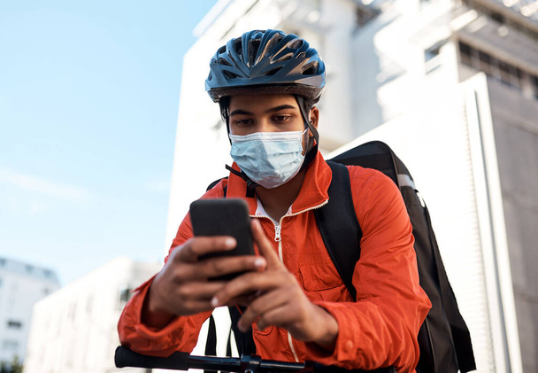 Seine Lieferungen steuert er mit seinem Smartphone. Aufnahme eines maskierten Mannes, der sein Handy benutzt, während er auf seinem Fahrrad für eine Lieferung unterwegs ist. - Foto, Bild