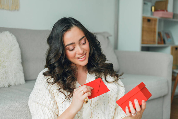 Emotional portrait of a 30 year old woman in knitted stylish cardigan. Young beautiful dark haired curly woman holding a red envelope greeting card on valentine's day. Cozy home. - 写真・画像