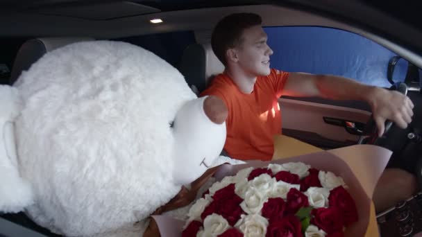 Szczęśliwy facet jedzie samochodem z bukietem kwiatów i wielkim misiaczkiem. Prezenty - Materiał filmowy, wideo