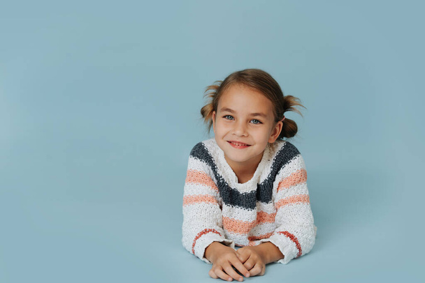 Portrait of a little girl in striped sweater lying on the floor over blue background. Leaning on her forearms. Hair in buns. - Foto, Bild