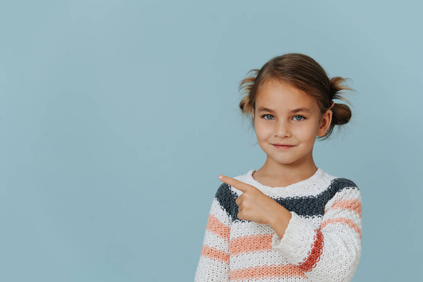 Grinning little girl in striped sweater pointing to side with index finger over blue background. - Foto, Imagen