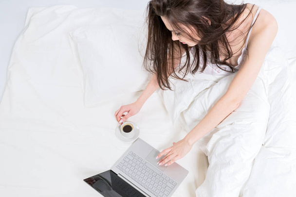 Una guapa morena está sentada en una cama blanca junto a un portátil y una taza de café. Buenos días. Trabajo desde casa. - Foto, imagen