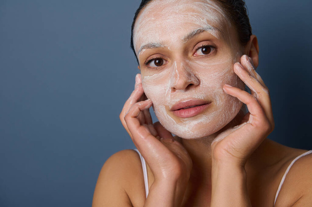 Zbliżenie młodej, ciemnoskórej kobiety, która robi sobie masaż twarzy, usuwa makijaż i czyści twarz złuszczającym kosmetykiem do oczyszczania twarzy, izolowanym na szarym tle - Zdjęcie, obraz