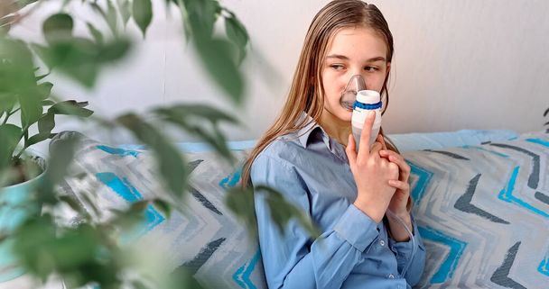 Teenage girl makes inhalation with a nebulizer equipment. Sick child holding inhalator in hand and breathes through an inhaler at home. Physical therapy for cold, flu and broncial asthma - Foto, Imagem