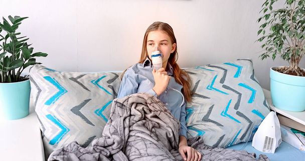 Teenage girl makes inhalation with a nebulizer equipment. Sick child holding inhalator in hand and breathes through an inhaler at home. Physical therapy for cold, flu and broncial asthma - Фото, изображение
