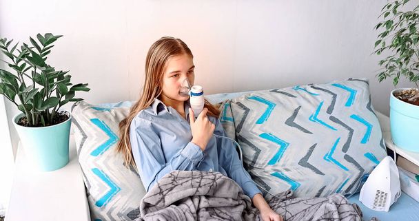 Teenage girl makes inhalation with a nebulizer equipment. Sick child holding inhalator in hand and breathes through an inhaler at home. Physical therapy for cold, flu and broncial asthma - Photo, image
