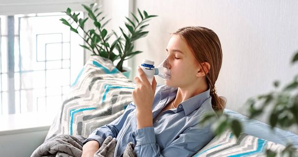 Teenage girl makes inhalation with a nebulizer equipment. Sick child holding inhalator in hand and breathes through an inhaler at home. Physical therapy for cold, flu and broncial asthma - 写真・画像