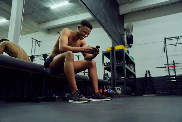 Young African American male looking at his phone to reply to an online message. Happy, mixed race male smiling at his phone while sitting down in the gym. High quality photo  - Foto, Imagem
