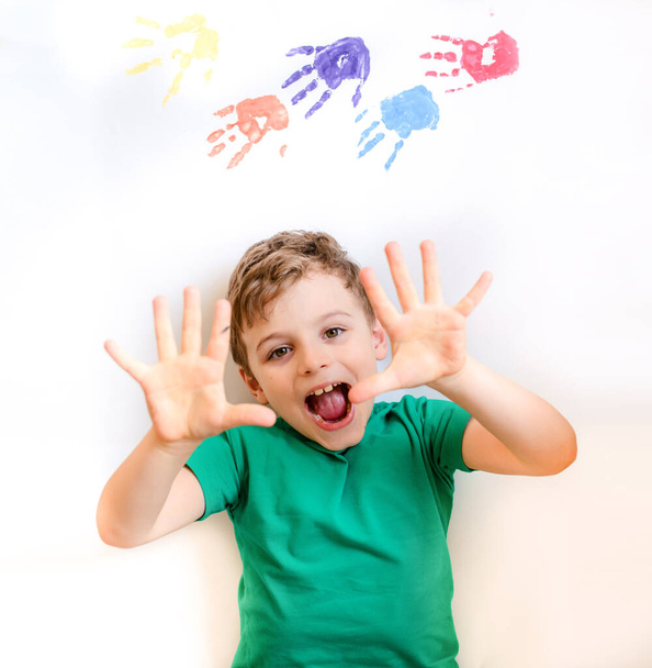 close-up kleine jongen glimlach en lachen naar de camera op witte achtergrond met kleur verf handen op de achtergrond. - Foto, afbeelding