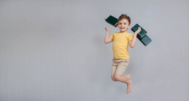 Education is funny and easy concept. Happy and smiling little boy child is standing on gray background and throw many books. Free space for text and logo on yellow t-shirt - Foto, afbeelding