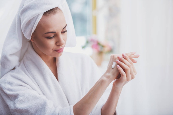 Beautiful young woman in a bathrobe with a towel on her head applying cream on her hands, standing in the bathroom - Foto, Imagem