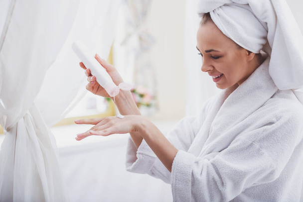 Hermosa joven en un albornoz con una toalla en la cabeza que aplica crema en la mano y sonríe mientras está de pie en el baño
 - Foto, imagen