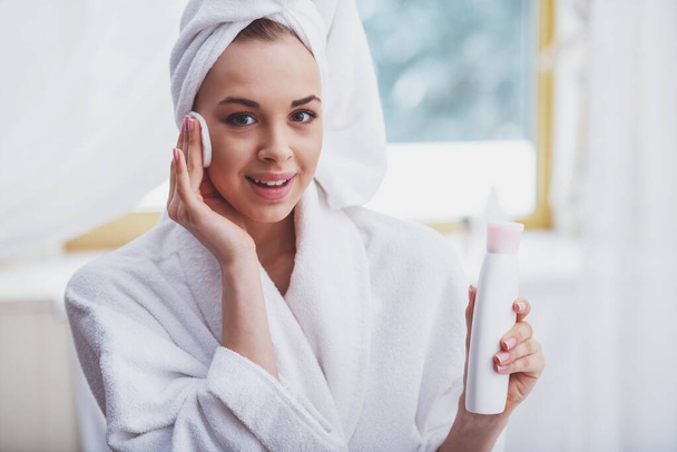 Красивая молодая женщина в халате с полотенцем на голове, держа бутылку с тонером и очищая лицо губкой, глядя в камеру и улыбаясь
 - Фото, изображение