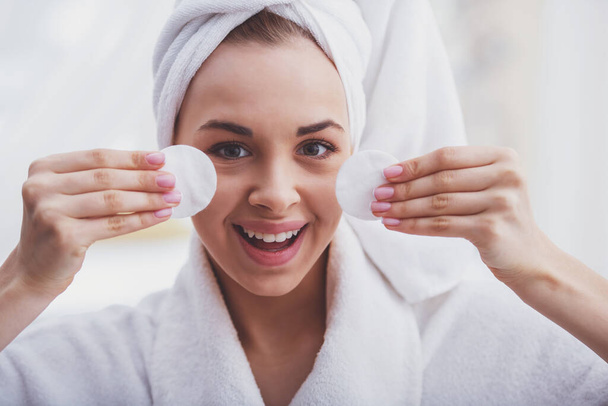 Retrato de una hermosa joven en un albornoz con una toalla en la cabeza sosteniendo esponjas en ambas manos, mirando a la cámara y sonriendo
 - Foto, Imagen