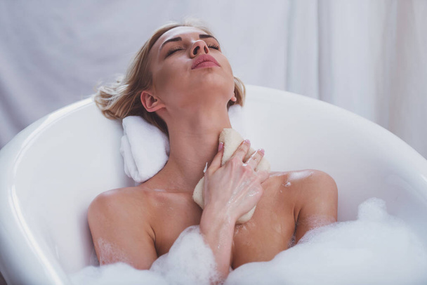 Молодая женщина моется, с помощью баста и наслаждается приятной ванной с пеной, лежа с закрытыми глазами
 - Фото, изображение