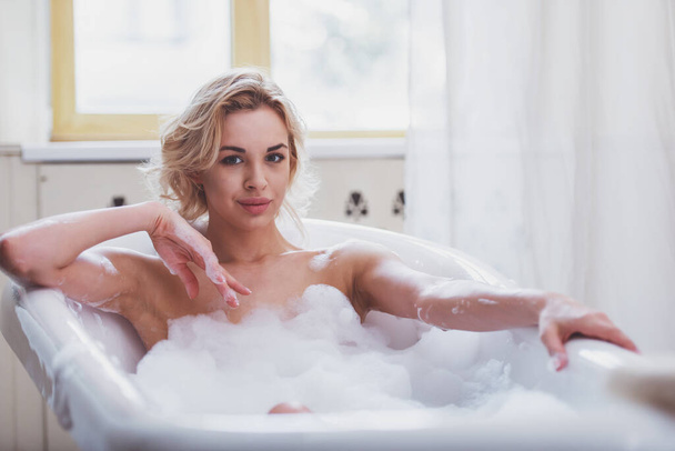 Hermosa joven rubia disfrutando de un baño agradable con espuma, mirando a la cámara y sonriendo
 - Foto, imagen